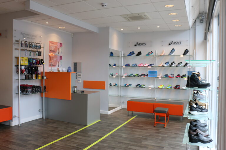 The inside of a shoe shop, with red accents and shoes lining the wall and a chair for shoe fitting.