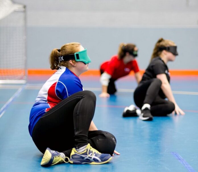 three teenagers sit in a sports hall with their eyes covered