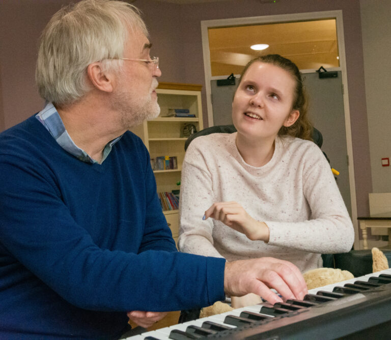 Dr Adam Ockleford, a man with gray hair and a blue jumper, plays the piano for Lily, who has juvenile dementia and is wearing a pink jumper