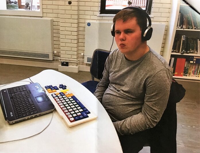 A photo of Ben, a young adult with BBS, sitting at a computer wearing headphones