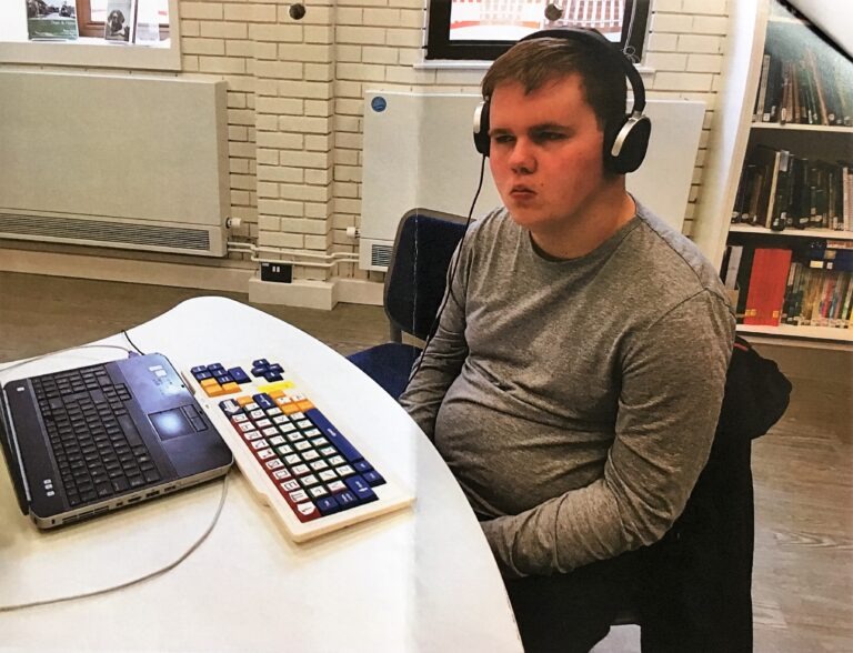A photo of Ben, a young adult with BBS, sitting at a computer wearing headphones