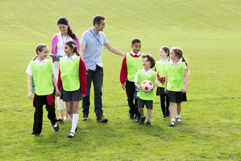 Group of school children wearing bibs and school uniform outside on the grass with their teacher. One of the boys is holding a soccor ball.